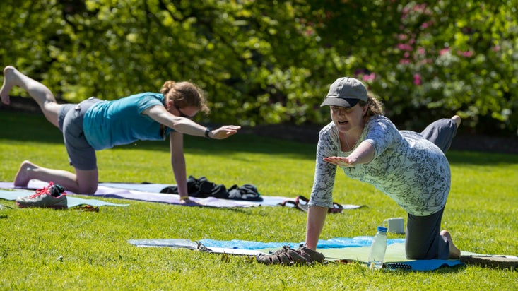 Two women are doing yoga positions, kneeling with one hand and the opposite foot in the air. They are on a green lawn surrounded by foliage.